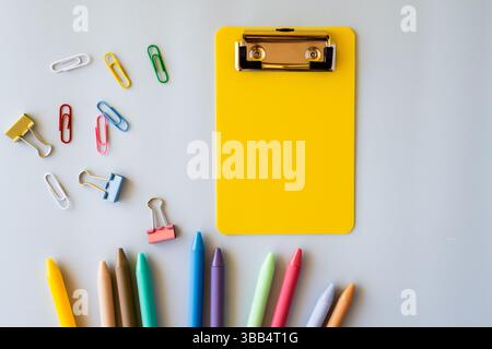Hellgelbes Klemmbrett mit verstreuten bunten Büroklammern und Stiften auf neutralem Hintergrund, flaches Design für Kreativität im Bildungsbereich Stockfoto