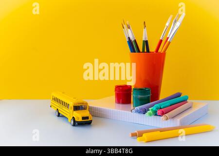 Kreative Schulbedarf Arrangement mit Pinseln farbige Stifte Schulbus und Notizbücher auf gelbem Hintergrund, die künstlerisches Lernen darstellen Stockfoto