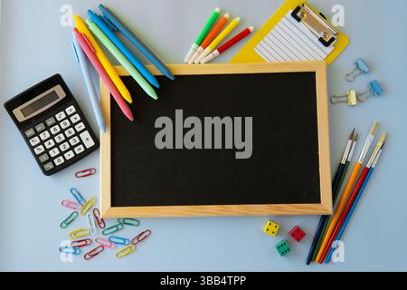 Farbenfrohe Back-to-School-Flatlay mit schwarzer Tafel Bright Pens Taschenrechner bürstet Clipboard Würfel und Büroklammern auf blauem Hintergrund kreativ Stockfoto