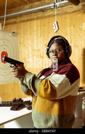 Porträt einer Frau im Schießstand im Innenbereich, die Waffen zum Testen anbietet und eine Schutzbrille trägt. Kunde mit Schutzausrüstung und Erfahrung mit Pistole Stockfoto