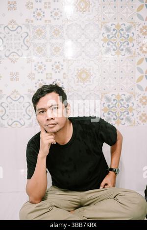 Ein asiatischer Mann sitzt nachdenklich drinnen und trägt legere Kleidung mit einem nachdenklichen Gesichtsausdruck. Stockfoto