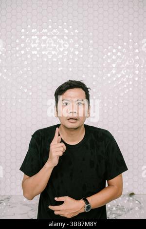 Ein ausdrucksstarker asiatischer Mann zeigt nach oben, ein nachdenklicher Blick auf sein Gesicht. Veranschaulicht die Konzepte von Ideen, Lösungen oder Bewusstseinsbildung. Stockfoto