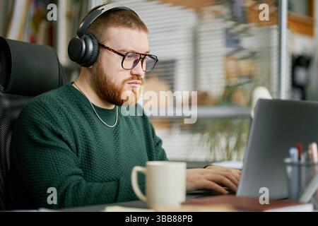 Porträt eines Mannes mit Kopfhörern, Brillen und Bart, der in modernen Büros am Laptop arbeitet. Fokussierter Ausdruck und professionelles Setting umgeben von Offi Stockfoto