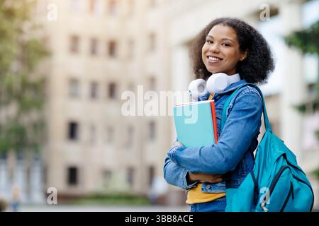 Fröhliche schwarze Schülerin mit Kopfhörern um den Hals, mit Notizbüchern und Rucksack Stockfoto