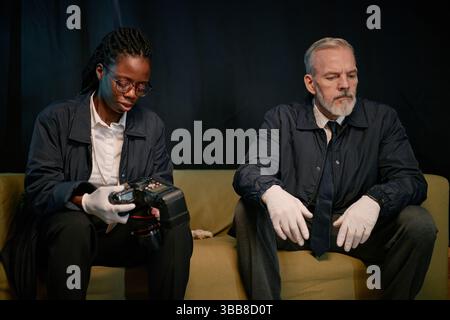 Afroamerikanische weibliche und ältere kaukasische männliche Kollegen arbeiten an der Untersuchung am Arbeitsplatz zusammen. Frau untersucht Geräte, während Mann s beobachtet Stockfoto
