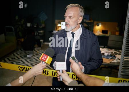 Interviewszene, die einen älteren Mann mit grauem Bart am Tatort im Büro zeigt, spricht und von Medienreportern mit Mikrofonen interviewt wird Stockfoto