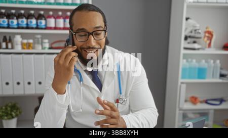 Junger Mann Arzt drinnen mit Bart, glücklich am Telefon in der modernen Klinik umgeben von medizinischen Geräten und Büroakten, Schaffung eines Berufs Stockfoto