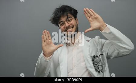 Junger hispanischer Mann mit Bart in Brille Lächeln und Gesten in einem weißen Mantel vor grauem Hintergrund, der Selbstvertrauen und Freundlichkeit zeigt. Stockfoto