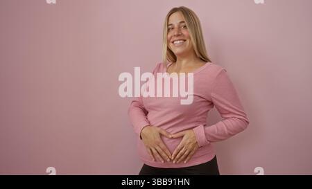 Schwangere Frau lächelt, hält ihren Bauch vor einem isolierten rosafarbenen Hintergrund und drückt Freude und Erwartung für ihre bevorstehende Babyreise aus. Stockfoto