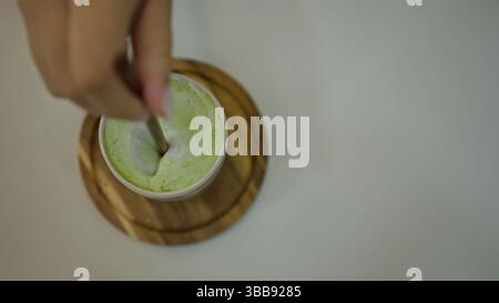 Matcha Latte mit Handrühren in einem Café mit cremiger Textur und leuchtender grüner Farbe auf einem Holzuntersetzer. Stockfoto