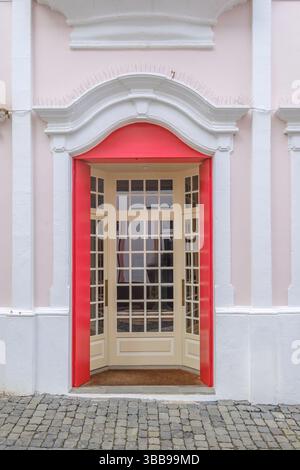 Verzierte Tür mit rotem Rahmen und rosafarbener Fassade, Terceira. Bezaubernde Aussicht auf eine elegante, cremefarbene, mehrverglaste Tür mit einem leuchtend roten Rahmen, in einem Stockfoto