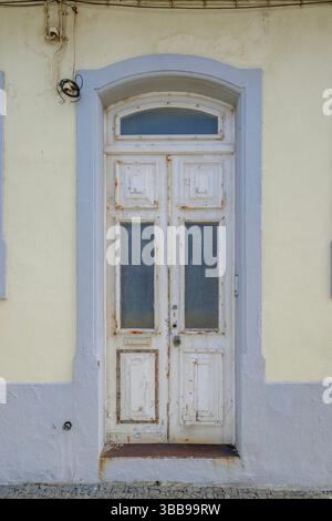 Verwitterte weiße Tür mit Bogenrahmen in Angra do Heroismo, Azoren. Rustikale Nahaufnahme einer alten, abgenutzten weißen Holzdoppeltür mit Glasscheiben und einer Stockfoto