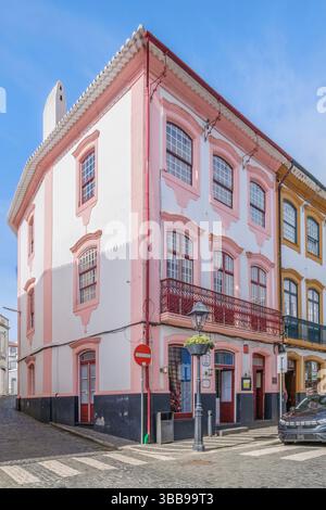 Eckgebäude mit Balkon in Angra do Heroismo, Azoren. Bezaubernde Aussicht auf ein dreistöckiges Gebäude an einer Ecke, weiße und rosa Fassade, verzierte Fenster, Stockfoto