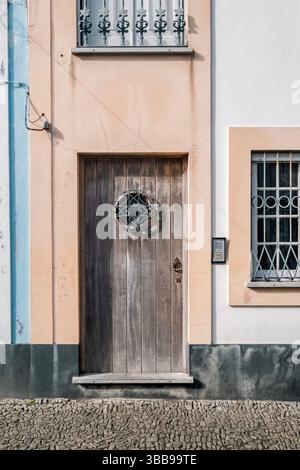 Rustikale Holztür mit rundem Fenster und schmiedeeisernen Details an der traditionellen Terceira-Fassade. Klassischer verwitterter Holzeingang auf pastellfarbenem Bu Stockfoto