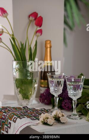 Stillleben mit einer Flasche Sekt, Kristallgläsern, roten Tulpen und Flieder in einer Vase auf einem Tisch mit traditioneller Stickerei Stockfoto