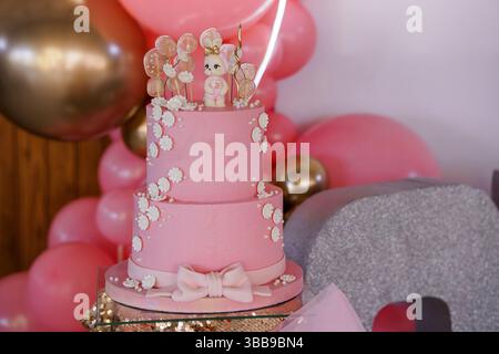 Ein bezaubernder rosafarbener zweistufiger Geburtstagskuchen mit einer niedlichen Hasenfigur und weißen Blumenakzenten, der vor einem festlichen Ballonbogen positioniert ist Stockfoto
