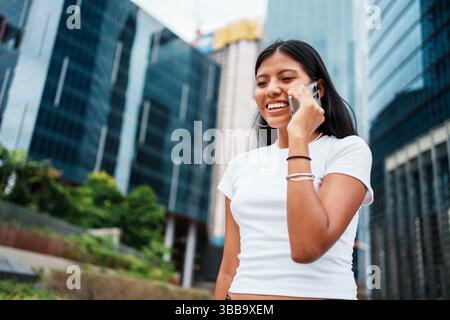 Eine lächelnde junge Frau, die sich in einem lebhaften Telefongespräch in einer lebhaften urbanen Umgebung verstrickt hat Stockfoto