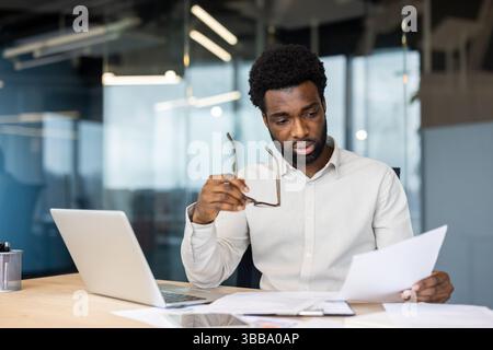 Ein fokussierter schwarzer Geschäftsmann analysiert Dokumente, während er seine Brille hält und fleißig an seinem Schreibtisch arbeitet. Er sitzt in einem Büro. Stockfoto