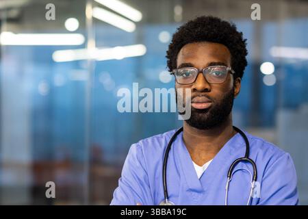 Ein gutaussehender, junger Arzt steht selbstbewusst in einer Arztpraxis. Seine Kleidung umfasst Peelings und ein Stethoskop. Das Büro ist im Hintergrund unscharf. Stockfoto