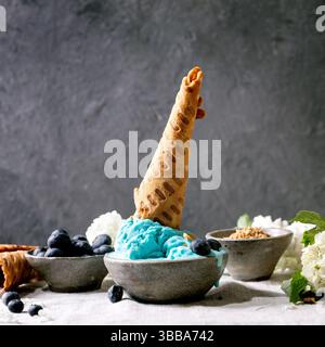 Hausgemachtes türkisfarbenes Eis mit Waffelkegel in Keramikschale. Heidelbeeren und weiße Blumen herum. Stehende graue Tischdecke aus Leinen. Trendig hausgemacht Stockfoto