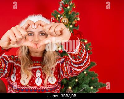 Nahaufnahme eines Weihnachtsmanns, der durch die Hände die Herzform schaut, steht über rotem Hintergrund in der Nähe des verzierten Weihnachtsbaums. Tragen Sie einen Weihnachtsmann-Hut, einen gemütlichen Pullover Stockfoto