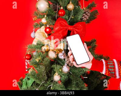 Weihnachtstelefon, Nahaufnahme von Hand, die Weihnachtstelefon hält. Leeres Smartphone mit weißem Bildschirm. Dekorierter Baumhintergrund, rote Wand. Stockfoto