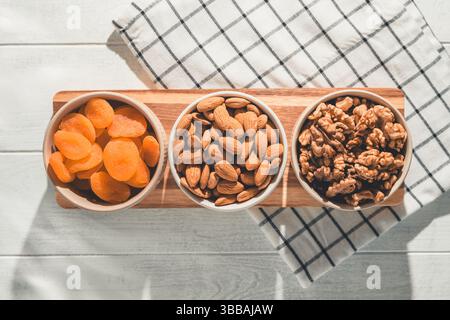 Drei Schüsseln mit gesunden Snacks, darunter getrocknete Aprikosen, Walnüsse und Mandeln, sind auf einem Holzbrett angeordnet und bieten eine nahrhafte und köstliche Leckerei Stockfoto