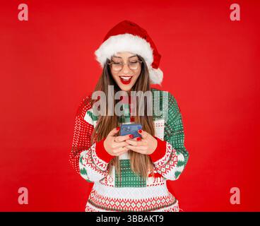 fröhlicher Spaß aufgeregt glückliche Frau in Santa Hut posiert halten in der Hand Handy benutzen. Trägt einen gestrickten Weihnachtspullover. Roter Hintergrund. Frohes neues Jahr Stockfoto