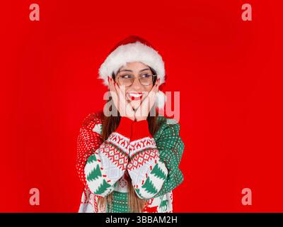 Kaukasische schockierte Frau. Überraschte brünette Weihnachtsmann in gestricktem Weihnachtspullover und Hut lächelnd, halten die Gesichter und schauen beiseite. Stockfoto