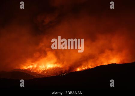 Nachts brennt ein gewaltiges Waldfeuer durch einen bewaldeten Hügel, wobei intensive Flammen und Rauch den dunklen Himmel in einer klimabedingten Katastrophe erhellen Stockfoto