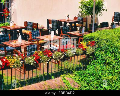 Charmante Terrasse im Freien mit Holztischen und -Stühlen, mit bunten Blumen und üppigem Grün, die eine gemütliche und einladende Atmosphäre schaffen, ideal zum Entspannen. Stockfoto