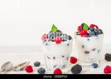 Griechischer Joghurt mit frischen Beeren, Hafer und Minze in Gläsern mit Löffeln auf weißem Hintergrund. Cremiger Joghurt mit Heidelbeere, Himbeere, brombeere Stockfoto