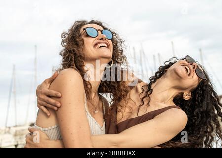 Nahaufnahme von zwei fröhlichen jungen Frauen mit lockigem Haar und Sonnenbrille, die sich auf einem Boot umarmen und lachen, mit Segelbootmasten im Hintergrund auf einem Stockfoto