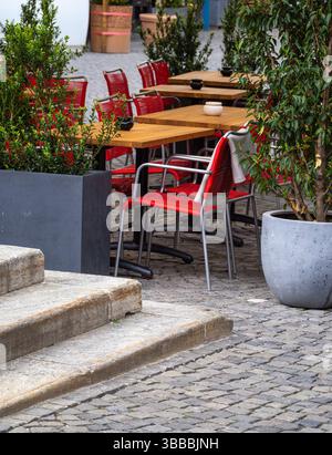 Ein gemütliches Café im Freien mit Holztischen und roten Stühlen, umgeben von Grün. Der Kopfsteinpflasterweg verleiht der Umgebung Charme, ideal für Stockfoto