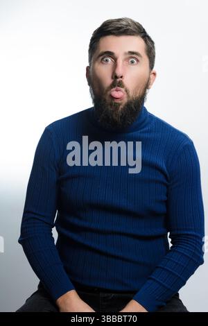 Ein bärtiger Mann in einem blauen Rollkragen streckt seine Zunge vor einem weißen Hintergrund aus. Sein spielerischer Ausdruck vermittelt Humor und Unbeschwertheit. Stockfoto