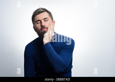 Ein bärtiger Mann in einem blauen Pullover hält seine Wange mit geschlossenen Augen vor einem einfachen Hintergrund. Sein Ausdruck deutet auf Unwohlsein oder mögliche Zahnschmerzen hin. Stockfoto