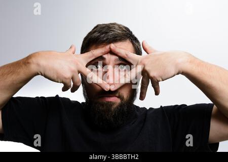 Ein bärtiger Mann, der ein schwarzes T-Shirt trägt, bildet mit seinen Fingern eine Dreiecksform um seine Augen, die Neugier oder Verspieltheit gegen einen leichten bac vermittelt Stockfoto