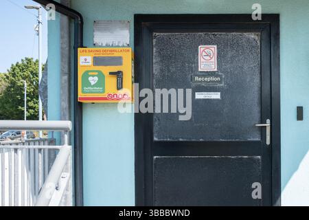 Glenarm, Nordirland - 14. Mai 2025 - lebensrettende Defibrillatormaschine an der Wand am Eingang zum Hafenamt im Hafen, Herzstillstand Stockfoto