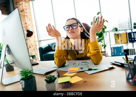 Junge Geschäftsfrau, die frustriert ist über ihren modernen Arbeitsplatz mit Dokumenten und Computer, was Stress symbolisiert Stockfoto