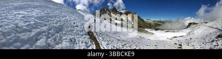 Panoramablick auf eine schneebedeckte Berglandschaft unter einem lebendigen Himmel, mit zerklüfteten Gipfeln und einem ruhigen Pfad, der die Schönheit der Natur und zeigt Stockfoto