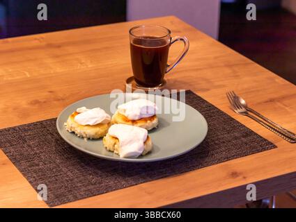 Traditioneller russischer Frühstückskäsekuchen oder Syrniki aus Hüttenkäse auf dem Teller neben einer Tasse Kaffee Stockfoto