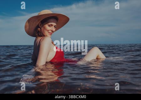Schöne junge Frau in rotem Badeanzug und Hut, die auf dem Meer im Wasser sitzt Stockfoto