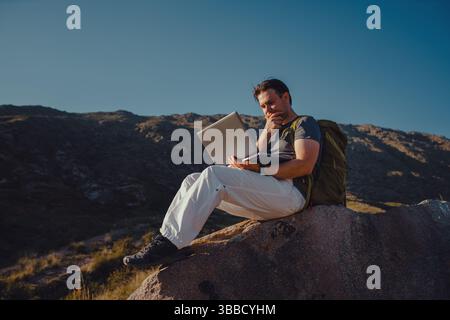 Ein junger Tourist mit Rucksack und Laptop sitzt auf dem Berg Stockfoto