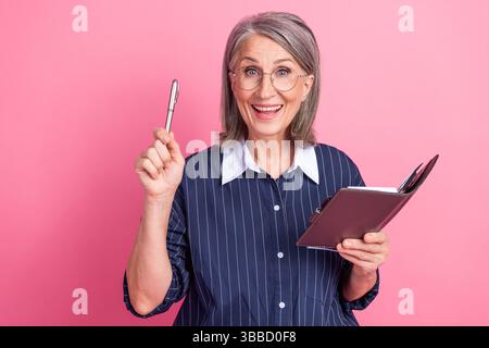 Lächelnde grauhaarige Frau, die ein Notizbuch und einen Stift hält, zeigt Aufregung vor einem rosafarbenen Hintergrund Stockfoto
