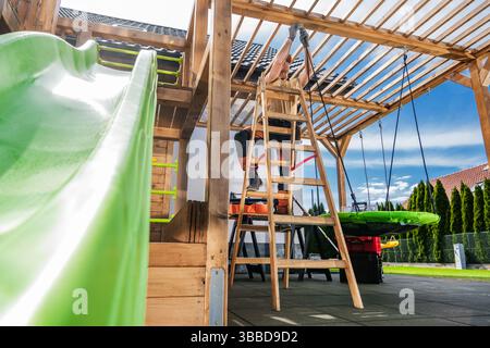 Ein Kind gleitet gerne auf einer hellgrünen Rutsche hinunter, während ein Elternteil in einem geräumigen Hinterhof daran arbeitet, eine Holzspielstruktur zu bauen. Die Sonne scheint b Stockfoto