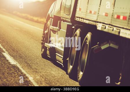 Ein großer Lkw fährt bei Sonnenuntergang über eine weitläufige Autobahn und wirft warmes goldenes Licht über die Landschaft. Der Fokus liegt auf den Hinterrädern, hoch Stockfoto