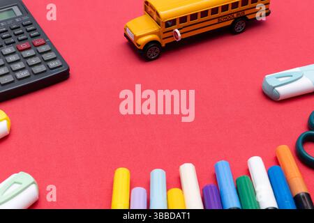 Verschiedene Schulutensilien, darunter ein Spielzeugbus, farbenfrohe Marker und Taschenrechner auf rotem Hintergrund Stockfoto