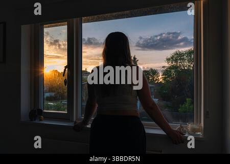 Eine junge Frau schaut aus ihrem Schlafzimmerfenster bei Sonnenuntergang Stockfoto