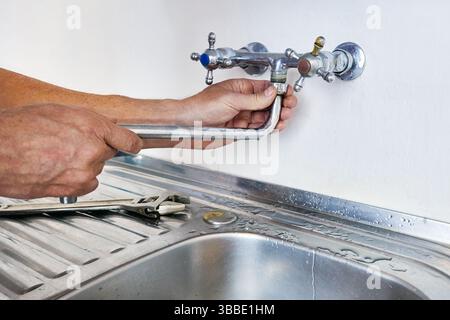Während der Reparatur des Wasserhahns montiert der Klempner den gebogenen Auslauf an den Doppelgriff über dem Edelstahlbecken, die Wasseranschlüsse sind in die Betonwand und den Schraubenschlüssel eingebettet Stockfoto