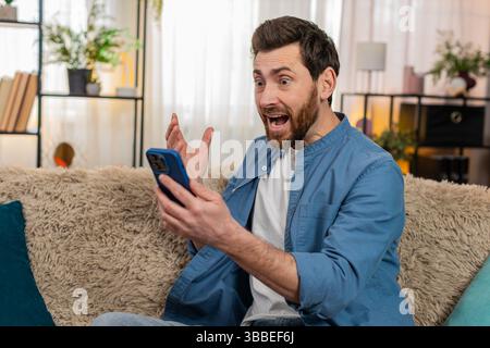 Junger erwachsener Mann, der zu Hause sitzt und soziale Medien auf dem Smartphone mit zielgerichtetem Ausdruck durchblättert. Der kaukasische Kerl auf dem Sofa hebt plötzlich überraschend die Augenbrauen, nachdem er unerwartete Online-Inhalte entdeckt hat Wow Stockfoto